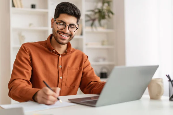 Hombre árabe sonriente en gafas usando laptop y tomando notas — Foto de Stock