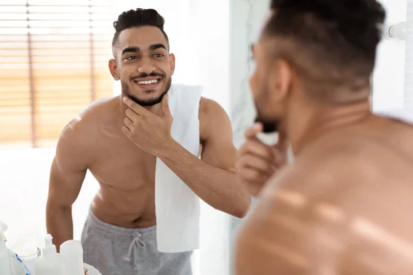 Cuidado de belleza masculino. Atractivo hombre árabe sin camisa mirando en espejo en el baño — Foto de Stock