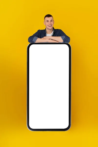 Chico feliz apoyado en la pantalla vacía del teléfono inteligente enorme, fondo amarillo — Foto de Stock