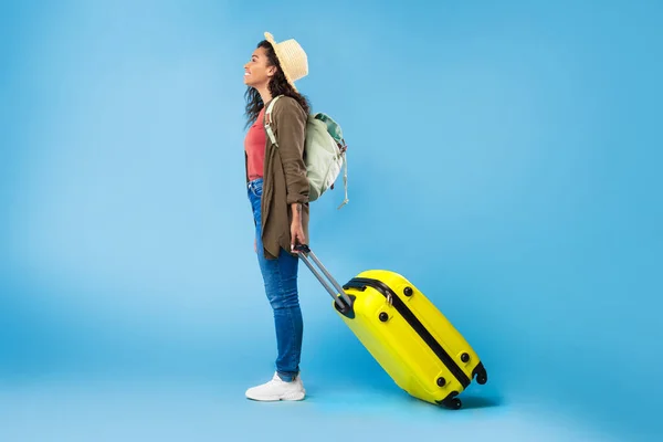 Vista laterale della giovane donna nera con zaino e valigia luminosa che va in vacanza, viaggia all'estero — Foto Stock