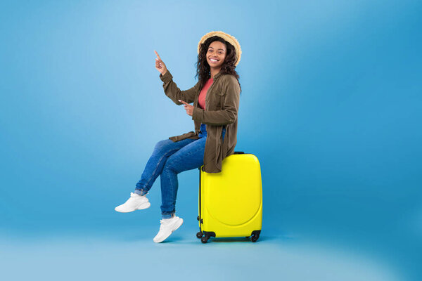 Happy young black woman sitting on bright suitcase and pointing at empty space for travel agency ad on blue background