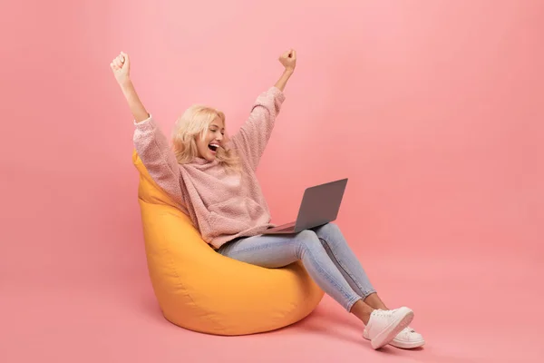 Senhora excitada fazendo gesto SIM, sentada em cadeira de saco de feijão com laptop e celebrando o sucesso, fundo rosa — Fotografia de Stock