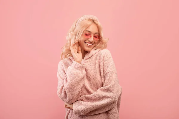 Señora bastante elegante mirando hacia abajo y sonriendo, con gafas de sol de colores y sudadera con capucha, posando sobre fondo rosa — Foto de Stock