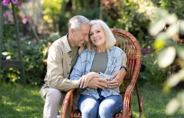 Milující starší manželé odpočívají na venkově, žena sedí v proutěném křesle a muž objímají ženu a usmívají se — Stock fotografie