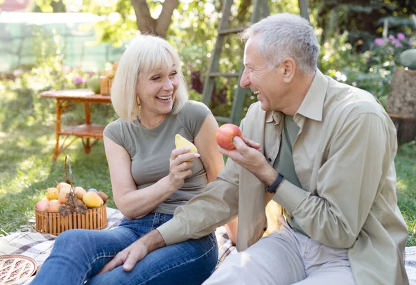 Сміються старші подружжя сидять на польовій траві і їдять фрукти, розмовляють і посміхаються, відпочивають на відкритому повітрі в своєму саду — стокове фото