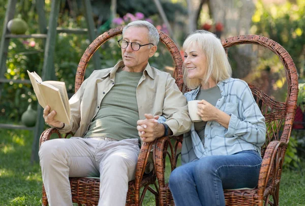 Loving elderly couple reading book together, sitting in wicker chairs and spending spring evening in their garden — Stock Photo, Image