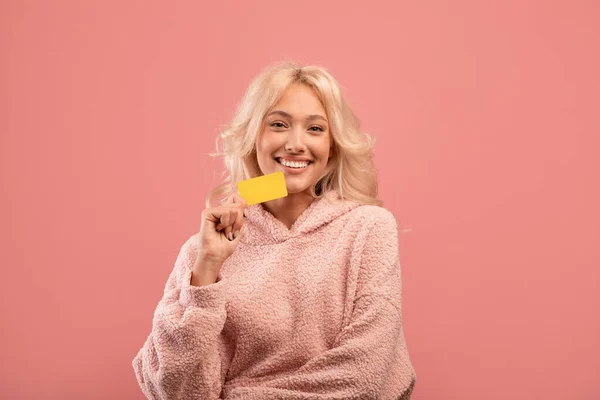 Koncepcja zakupów bez gotówki. Wesoła kobieta trzymająca kartę kredytową i uśmiechnięta do kamery, stojąca na różowym tle — Zdjęcie stockowe