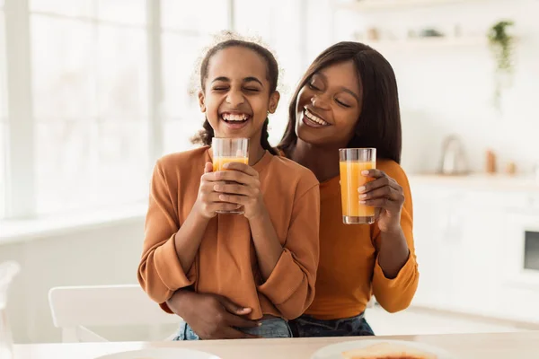 Negro mami e hija bebiendo naranja jugo riendo en cocina — Foto de Stock