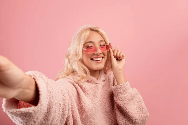 Mujer alegre y elegante haciendo selfie, con gafas de colores y sudadera con capucha, sonriendo a la cámara sobre fondo rosa — Foto de Stock