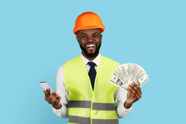 Trabalhador de construção feliz com dinheiro em dinheiro e Smartphone posando sobre fundo azul — Fotografia de Stock