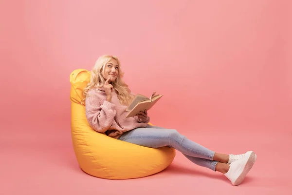 Snící dáma čtení knihy, zatímco sedí v pohodlné beanbag židle nad růžovým pozadím, kopírovat prostor — Stock fotografie