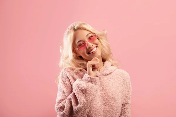 Jovencita feliz tocando la barbilla y sonriendo a la cámara, con elegantes gafas de sol de colores y sudadera con capucha, fondo rosa — Foto de Stock