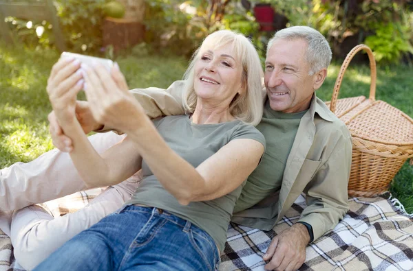 Glada äldre makar video ringer på smartphone samtidigt ha picknick och vila i trädgården på varma vårdagen — Stockfoto
