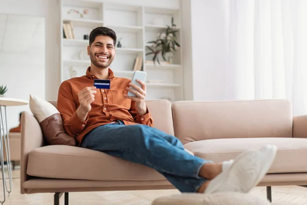 Sonriente chico árabe usando el teléfono y la tarjeta de crédito en casa — Foto de Stock