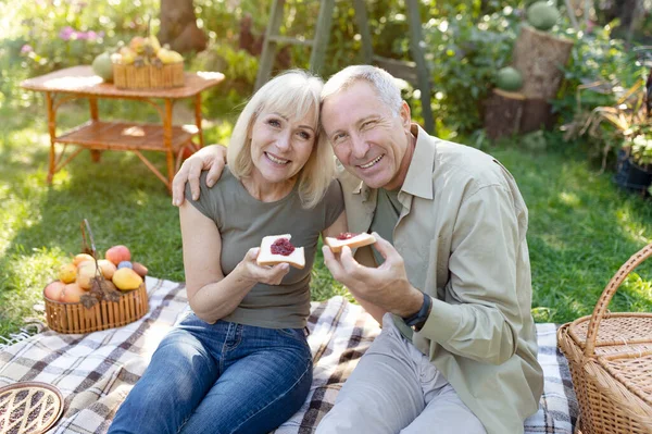 Kochający starsi małżonkowie urządzający piknik, siedzący na kocu w ogrodzie i jedzący tosty z dżemem, uśmiechający się do kamery — Zdjęcie stockowe