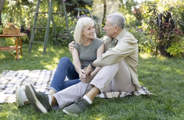 Szczęśliwa kochająca się para seniorów urządzająca piknik w ogrodzie, obejmująca się i uśmiechająca do siebie, siedząca razem na świeżym powietrzu — Zdjęcie stockowe