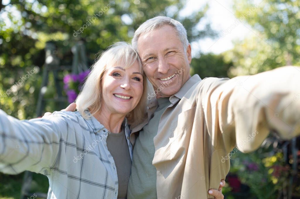 Happy senior spouses making selfie, hugging, smiling to camera, spending time together in their garden outdoors