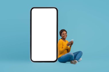 Elinde Akıllı Telefonla Büyük Boş Telefonun yanında oturan Mutlu Siyah Kadın