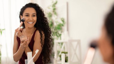 Neşeli genç bir kadın fırçayla toz sürüyor, evde aynanın önünde dekoratif makyaj yapıyor, boşluğu kopyalıyor.