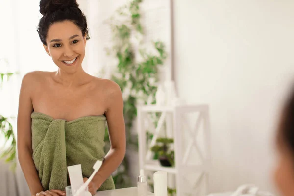 Περιποίηση ομορφιάς. Νεαρή όμορφη γυναίκα με πετσέτα κοιτάζει τον εαυτό της στον καθρέφτη και χαμογελά στο σπίτι, αντιγράφει χώρο — Φωτογραφία Αρχείου