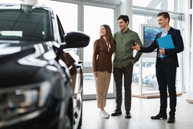 Otomobil galerisindeki genç çifte yeni otomobil gösteren mutlu araba satıcısı.