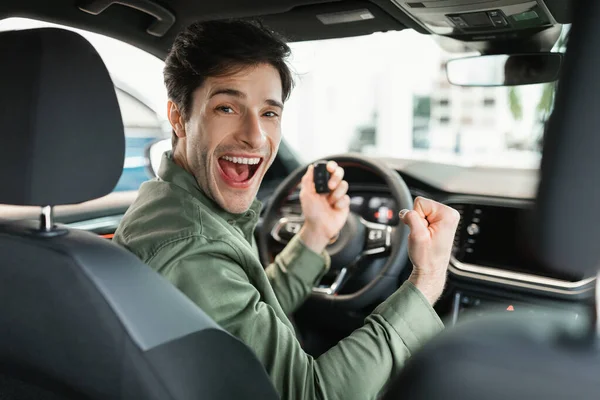 Caucasien excité gars tenant la clé de voiture et gestuelle OUI, assis dans une nouvelle voiture au magasin concessionnaire — Photo