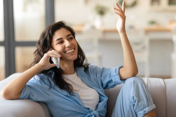 Joven morena alegre sentada en el sofá, conversando por teléfono móvil en casa, espacio para copiar — Foto de Stock