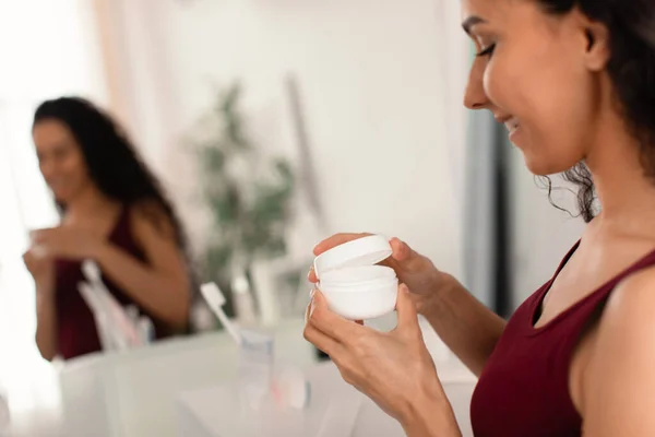 Mujer joven feliz con frasco de crema facial de pie delante del espejo, la aplicación de crema hidratante en casa, espacio para copiar —  Fotos de Stock