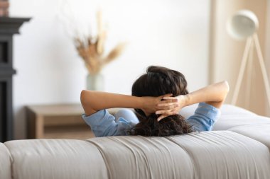 Genç esmer kadının arkası. Elleri başının arkasında kanepede oturuyor. Oturma odasında dinleniyor.