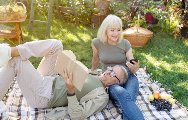 Отставные супруги отдыхают в саду, женщина пользуется смартфоном, а мужчина читает книгу, лежа на коленях у жен