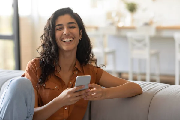 Giovane donna felice seduta sul divano in soggiorno, usando smartphone, chattando con un amico, comunicando online — Foto Stock