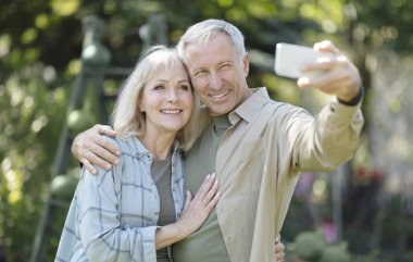 Mutlu yaşlı çift bahçede birlikte vakit geçiriyor, selfie çekiyor ve cep telefonu kameralarına gülümsüyor.