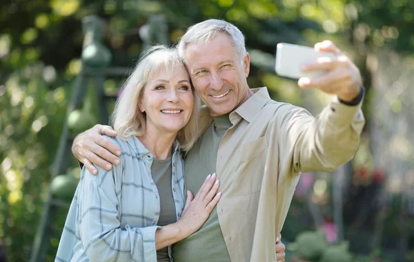 Щаслива літня пара проводить час у своєму саду разом, приймаючи селфі та посміхаючись у камері мобільного телефону — стокове фото