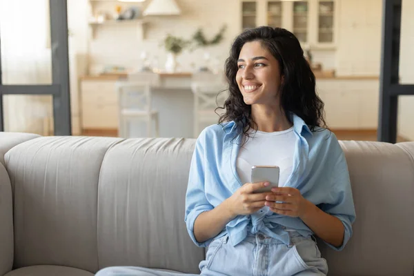 スマートフォンを自宅のソファに座っている若い女性の笑顔の肖像画、モバイルデバイスを使用して、コピースペース — ストック写真