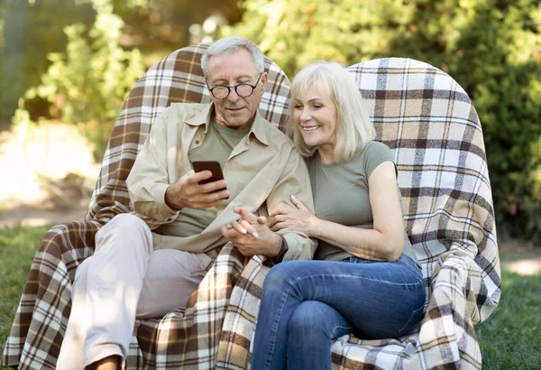 Glückliche ältere Eheleute, die es sich in Korbsesseln und mit dem Smartphone gemütlich machen und den warmen Frühlingsabend im Freien genießen — Stockfoto
