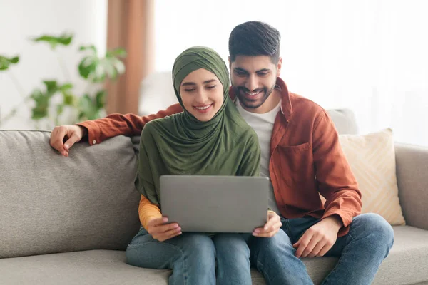 Coppia musulmana allegra che utilizza il computer portatile che naviga su Internet seduto a casa — Foto Stock