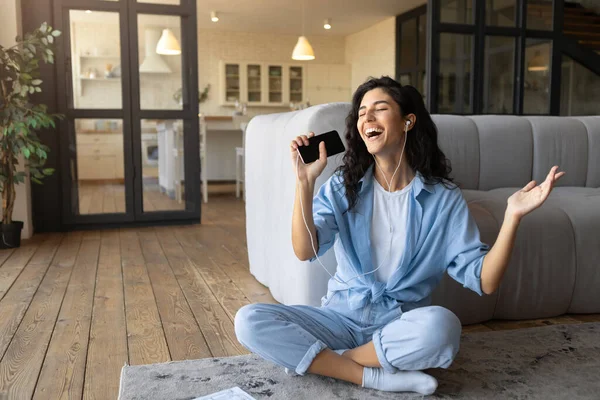 Jovencita alegre en auriculares escuchando música, cantando y usando smartphone como micrófono, sentada en el suelo en casa — Foto de Stock