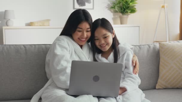 Ung glad asiatisk mamma och hennes lilla dotter bär badrockar tittar på digital-tv på bärbar dator, webb surfa tillsammans — Stockvideo