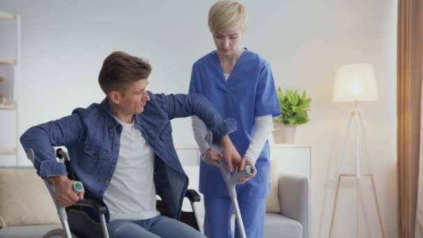 Первые шаги после аварии. Человек с инвалидностью встает с инвалидного кресла с помощью хрустов и реабилитатолога — стоковое видео