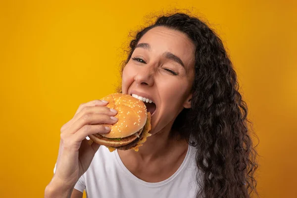 Com fome latina senhora segurando hambúrguer mordendo sanduíche no estúdio — Fotografia de Stock