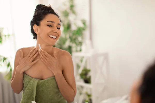 Perfect healthy skin concept. Pretty young woman in towel applying moisturizing cream on her neck near mirror at home — Stock Photo, Image