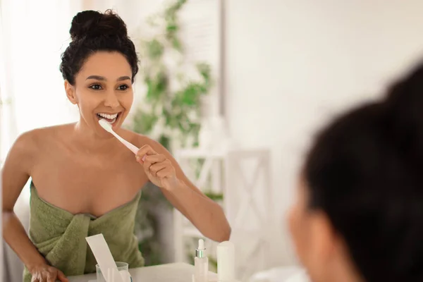 Bella signorina in asciugamano lavarsi i denti con spazzolino da denti vicino specchio a casa, copiare spazio. Concetto di igiene dentale — Foto Stock