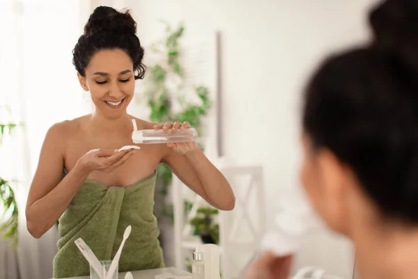 Mulher milenar bonita vestindo toalha após o banho, refrescando sua pele com loção ou tônico na frente do espelho no banheiro — Fotografia de Stock