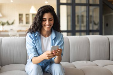 Genç esmer kadın koltukta cep telefonu kullanıyor, e-postaları kontrol ediyor, evde görüntülü sohbet ediyor, uzayı kopyalıyor.