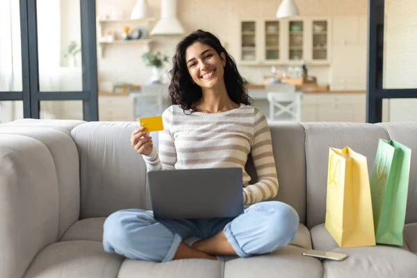 Jovencita sonriente sentada con las piernas cruzadas en el sofá, rodeada de bolsas de regalo, utilizando el ordenador portátil y la tarjeta de crédito en casa — Foto de Stock
