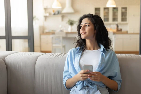 Giovane donna sognante seduta sul divano con smartphone, navigazione web, chat online, controllo dei social media a casa — Foto Stock