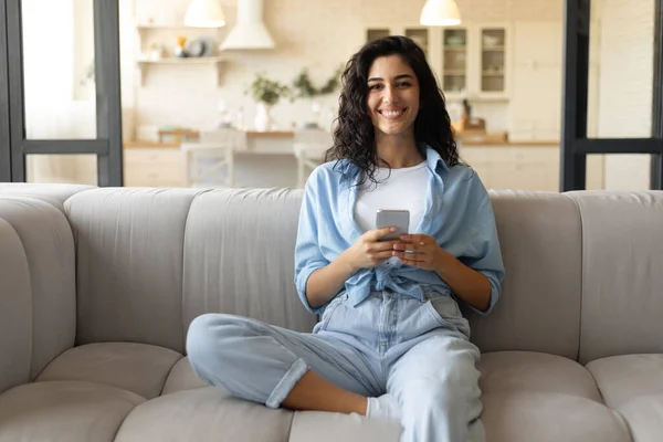 Happy young woman sitting on couch in living room and using cellphone, checking new mobile application at home — Stock Photo, Image