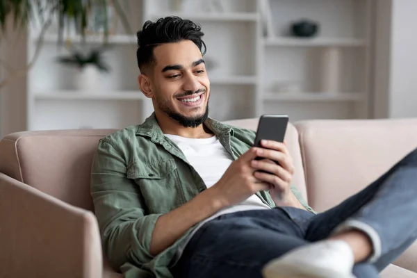 Retrato de chico árabe joven guapo relajante con teléfono inteligente en casa, —  Fotos de Stock