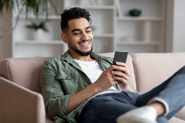 Retrato de bonito jovem árabe cara relaxante com smartphone em casa, — Fotografia de Stock