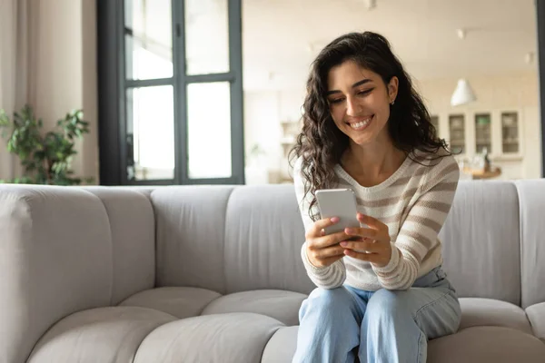 Millennial donna caucasica utilizzando smartphone mentre seduto sul divano, controllare i messaggi, avere videochiamata online a casa — Foto Stock